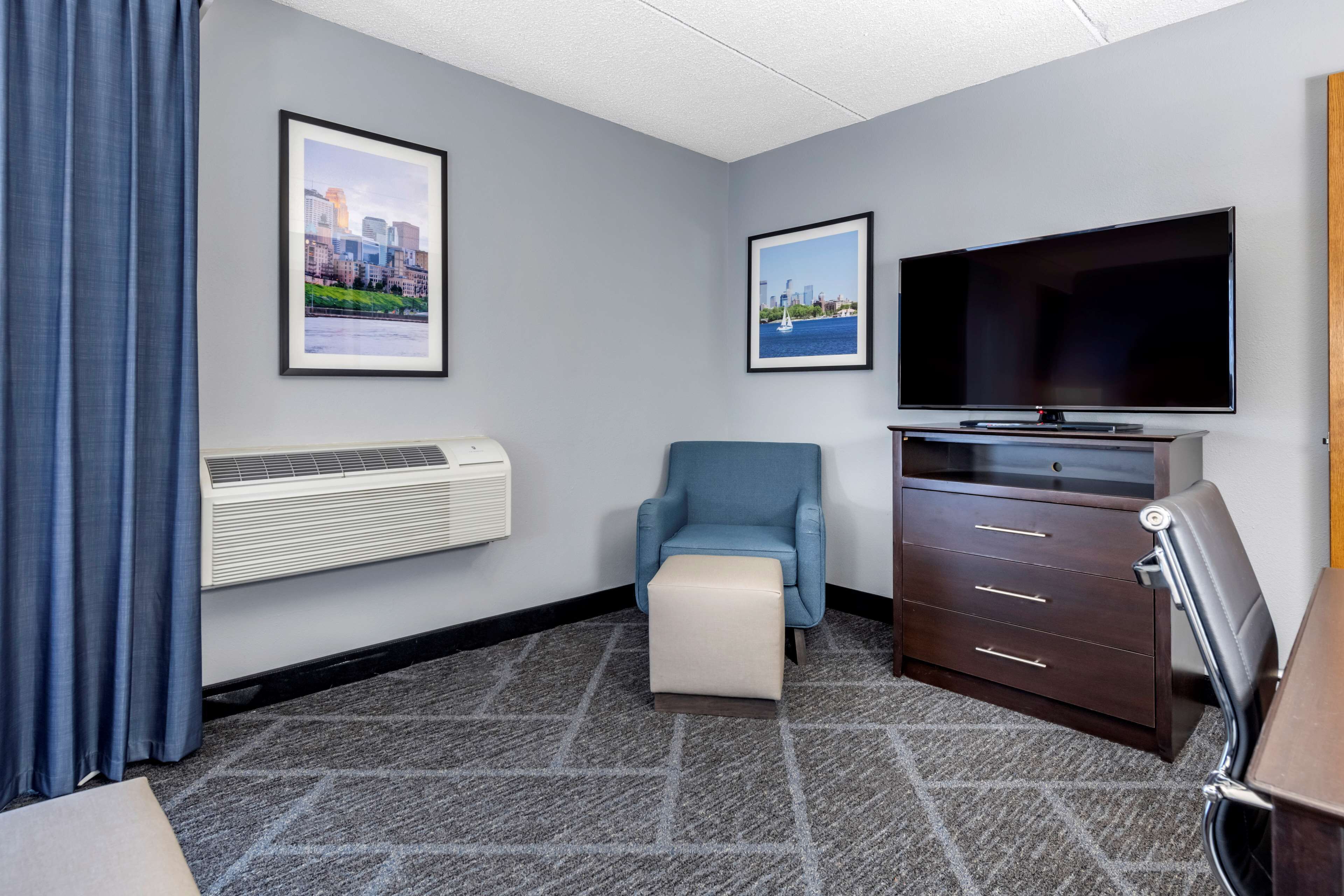 A hotel room with a large flat-screen TV, a blue armchair, a small ottoman, a dark wooden dresser, and two framed cityscape pictures on the wall.