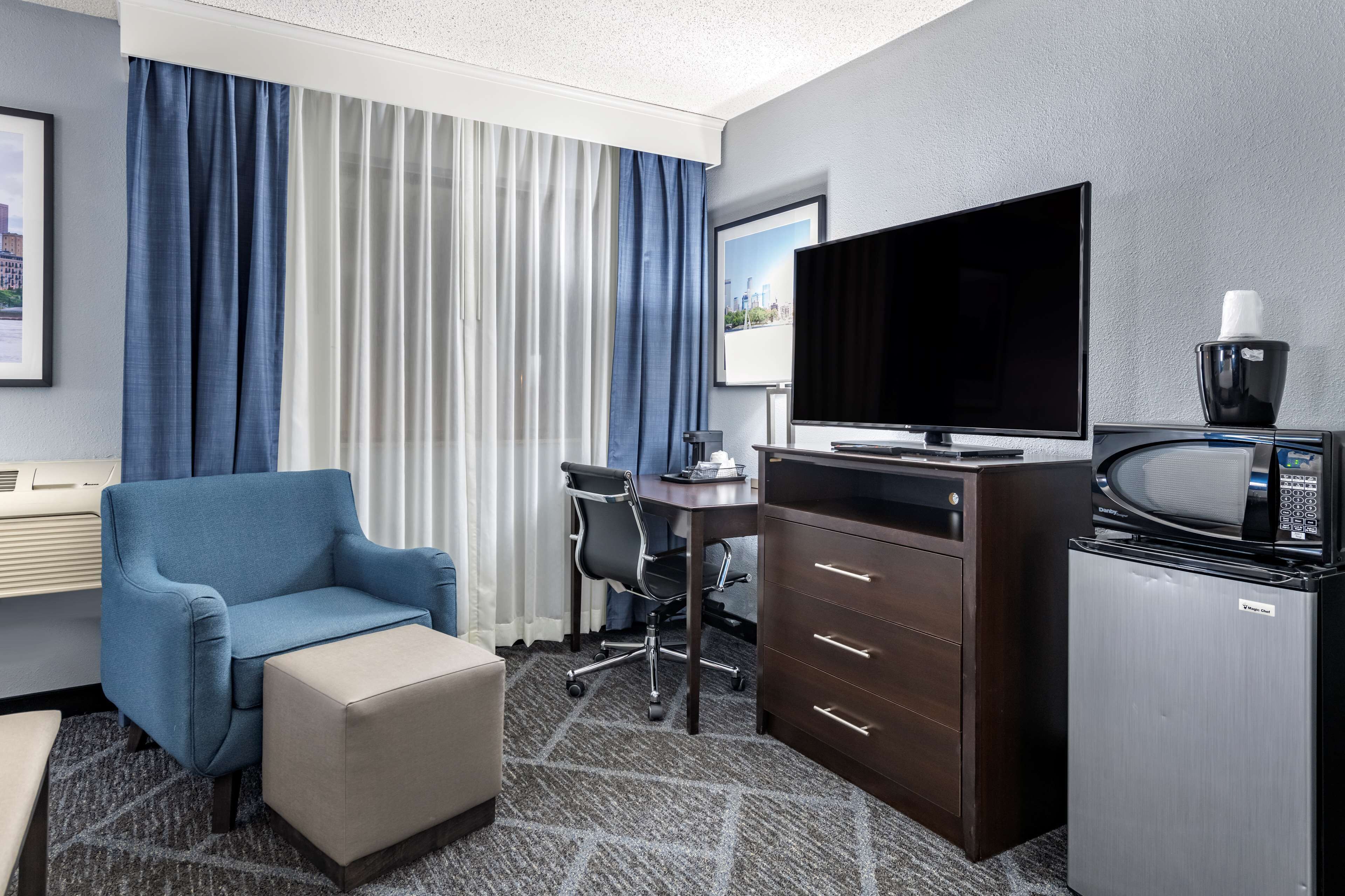 A hotel room with a blue armchair, a desk with a computer, a flat-screen TV, a microwave, and a refrigerator.