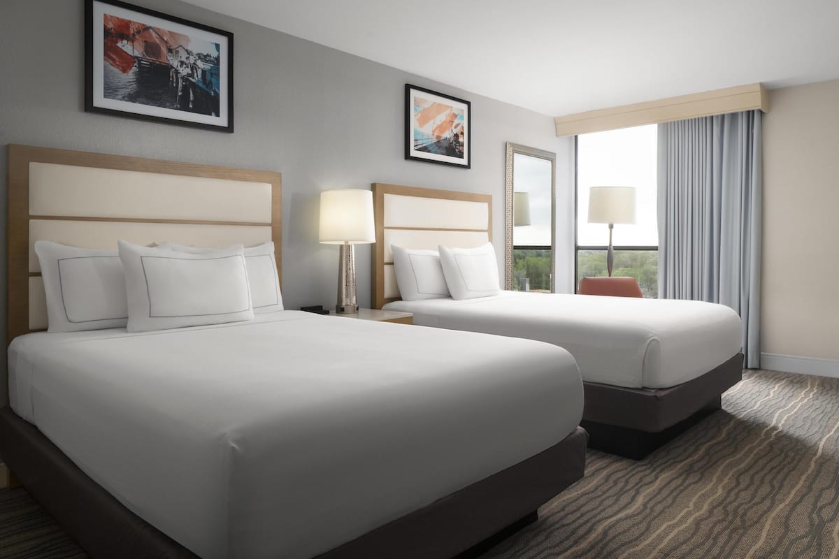Quarto de hotel com duas camas, uma janela grande com cortinas e duas fotografias emolduradas na parede.
