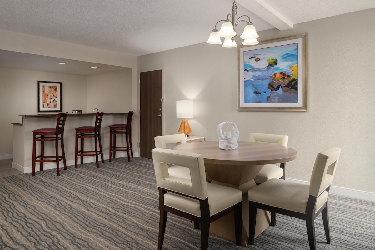 Quarto de hotel com área de jantar, mesa redonda e quatro cadeiras, pequena cozinha com bancada e banquetas, quadro decorativo na parede e tapete estampado.