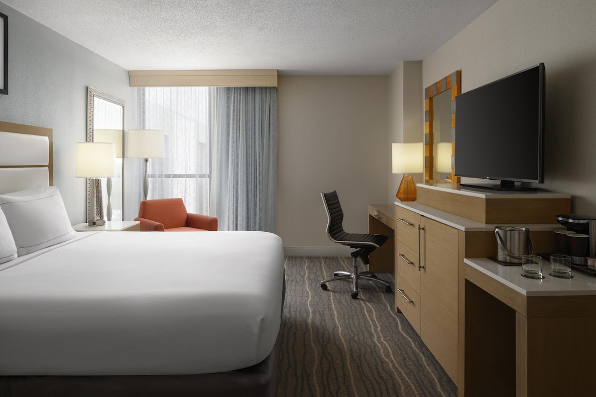 Quarto de hotel com uma cama grande, uma mesa com televisão, uma cadeira e uma luminária.