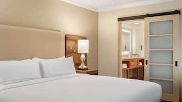 A hotel room with a large bed, a nightstand with a lamp, a wooden headboard, a sliding glass door to a bathroom, and a wooden dresser with a mirror.