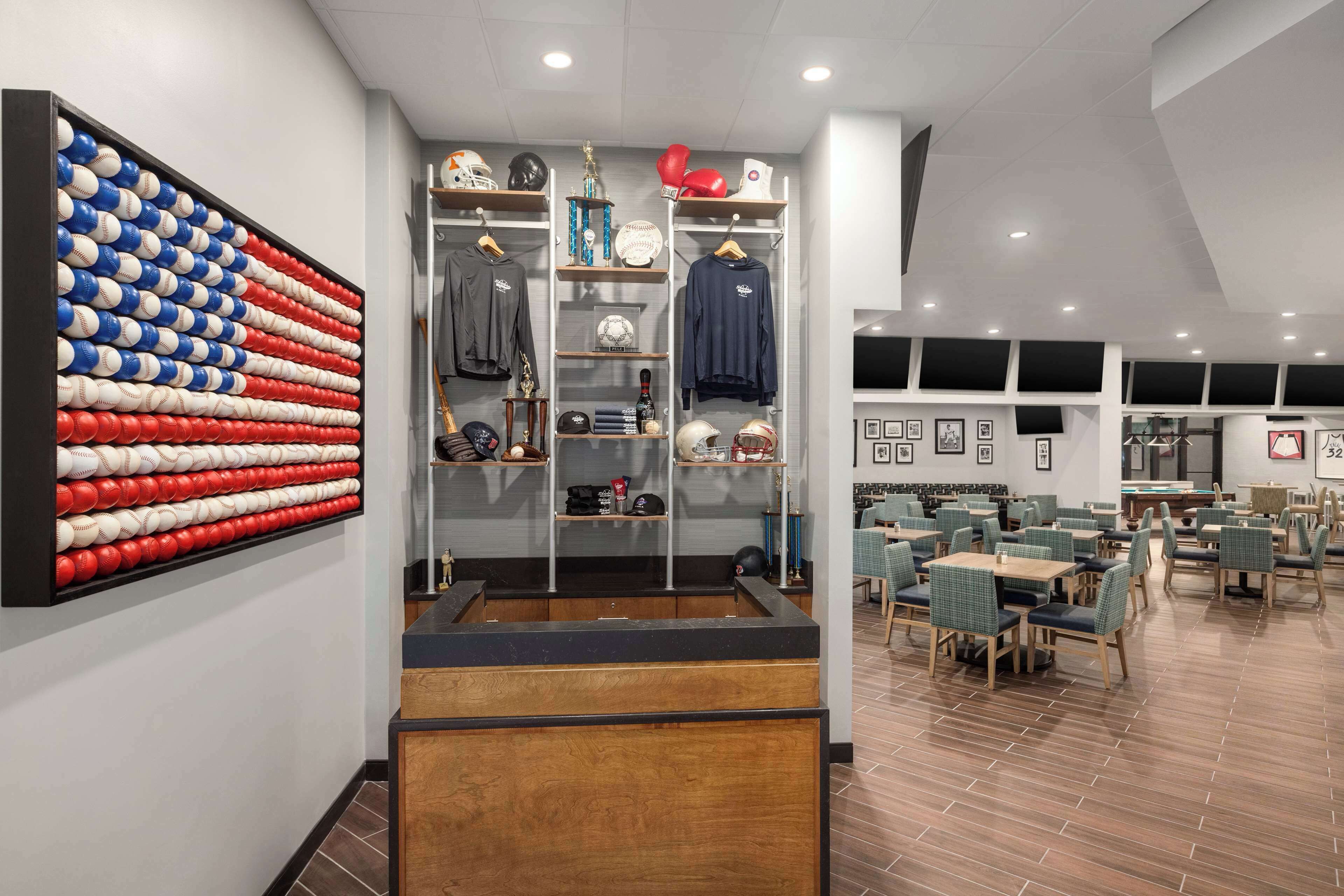 Un hall d’hôtel moderne agrémenté d’une œuvre d’art murale sur le thème du baseball, d’un espace de détente central avec des tables et des chaises, et de divers objets de collection liés au sport, exposés sur des étagères.