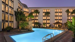 A swimming pool with a stone wall and palm trees, surrounded by a building with large windows and outdoor seating. - Doubletree by Hilton Fort Myers at Bell Tower Shops (Fort Myers)