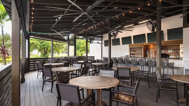 An outdoor patio area with round tables and chairs, ceiling fans, and a bar area with shelves of bottles. - Doubletree by Hilton Fort Myers at Bell Tower Shops (Fort Myers)