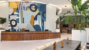 A modern reception area with a large abstract mural, a wooden reception desk, and a seating area with a long table and chairs. - Doubletree by Hilton Fort Myers at Bell Tower Shops (Fort Myers)