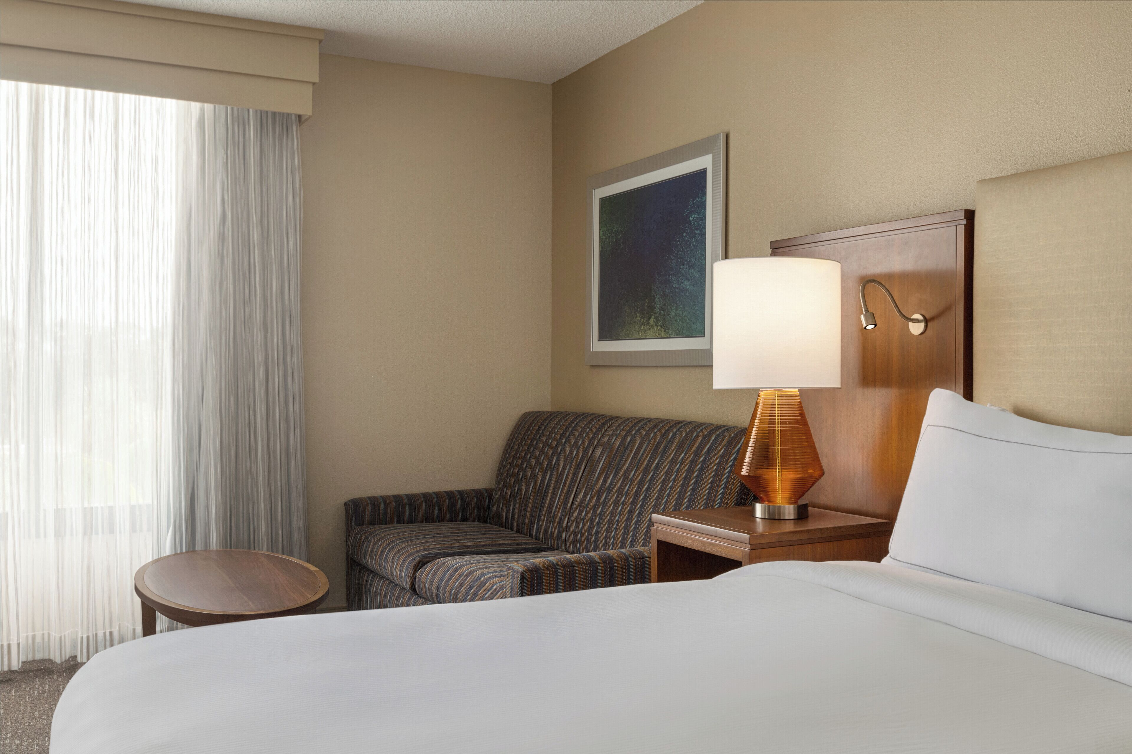 A hotel room with a bed, a striped armchair, a small table, a lamp, a painting on the wall, and a window with sheer curtains.