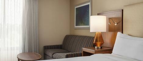 A hotel room with a bed, a striped armchair, a small table, a lamp, a painting on the wall, and a window with sheer curtains.