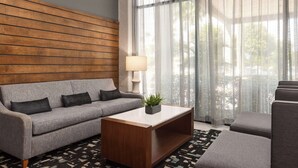 A modern living room with a grey sofa, a wooden accent wall, a coffee table with a plant, and sheer curtains allowing natural light. - Doubletree by Hilton Fort Myers at Bell Tower Shops (Fort Myers)
