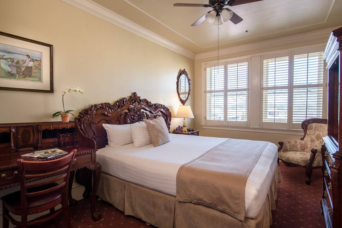 A spacious bedroom with a large bed, a desk, a chair, and a window with blinds.