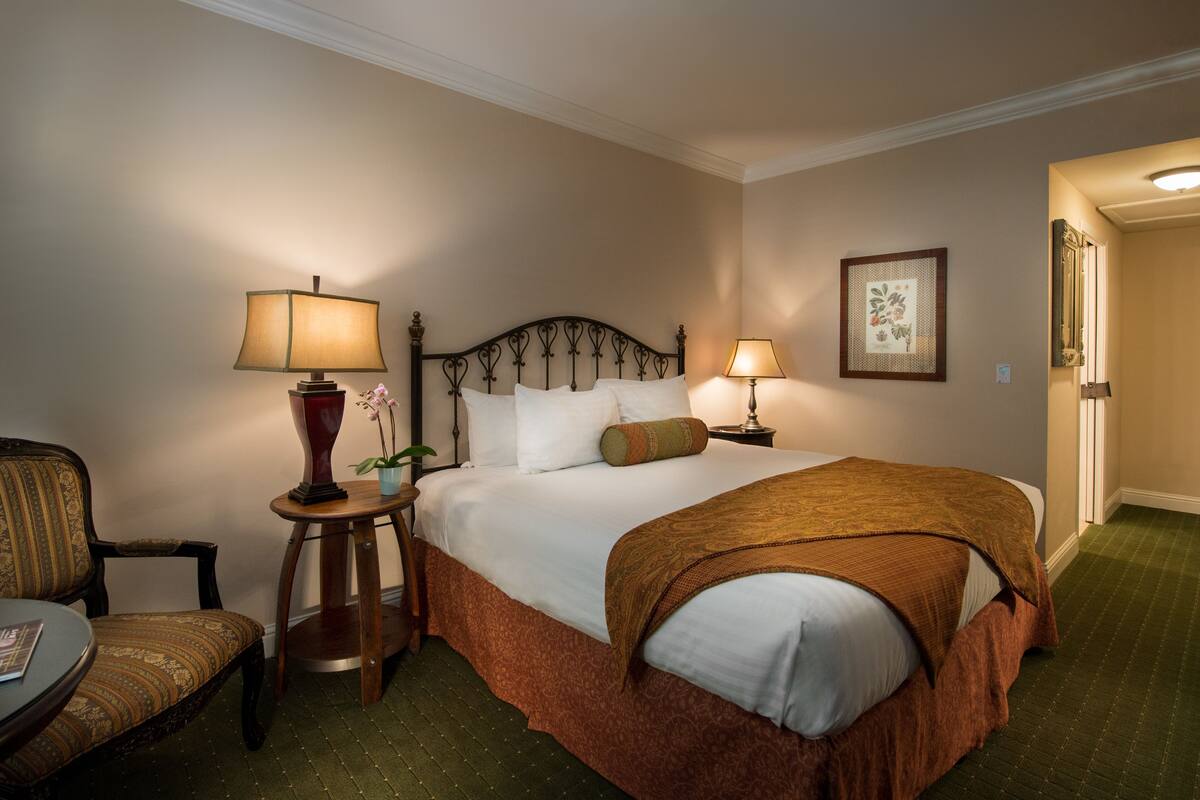 A hotel room with a bed, two bedside lamps, a chair, a small table, and a framed picture on the wall.