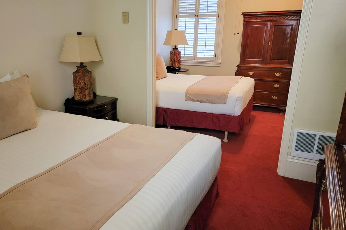 A hotel room with two beds, a nightstand with a lamp, a dresser, and a window with blinds.