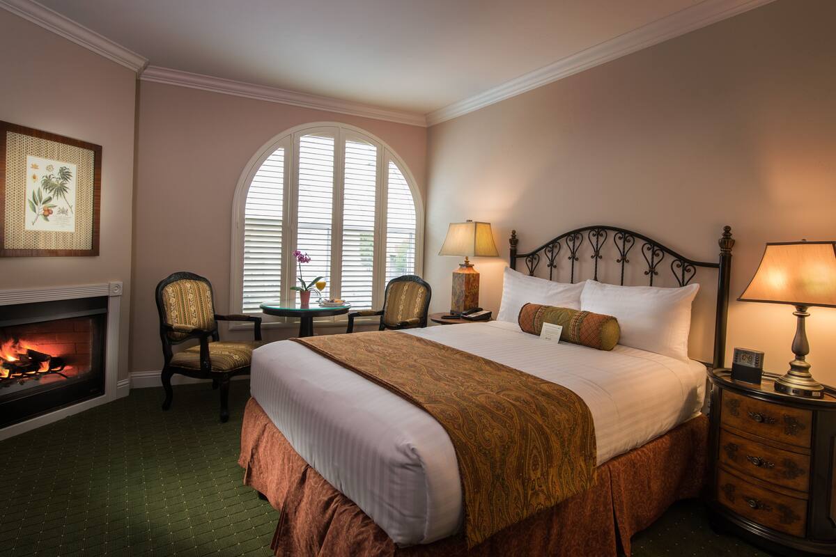 A hotel room with a large bed, a fireplace, a window with blinds, a chair, and a nightstand with a lamp.