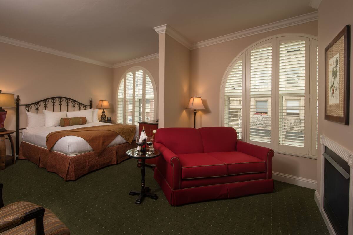 A hotel room with a bed, a red sofa, two lamps, and large arched windows.