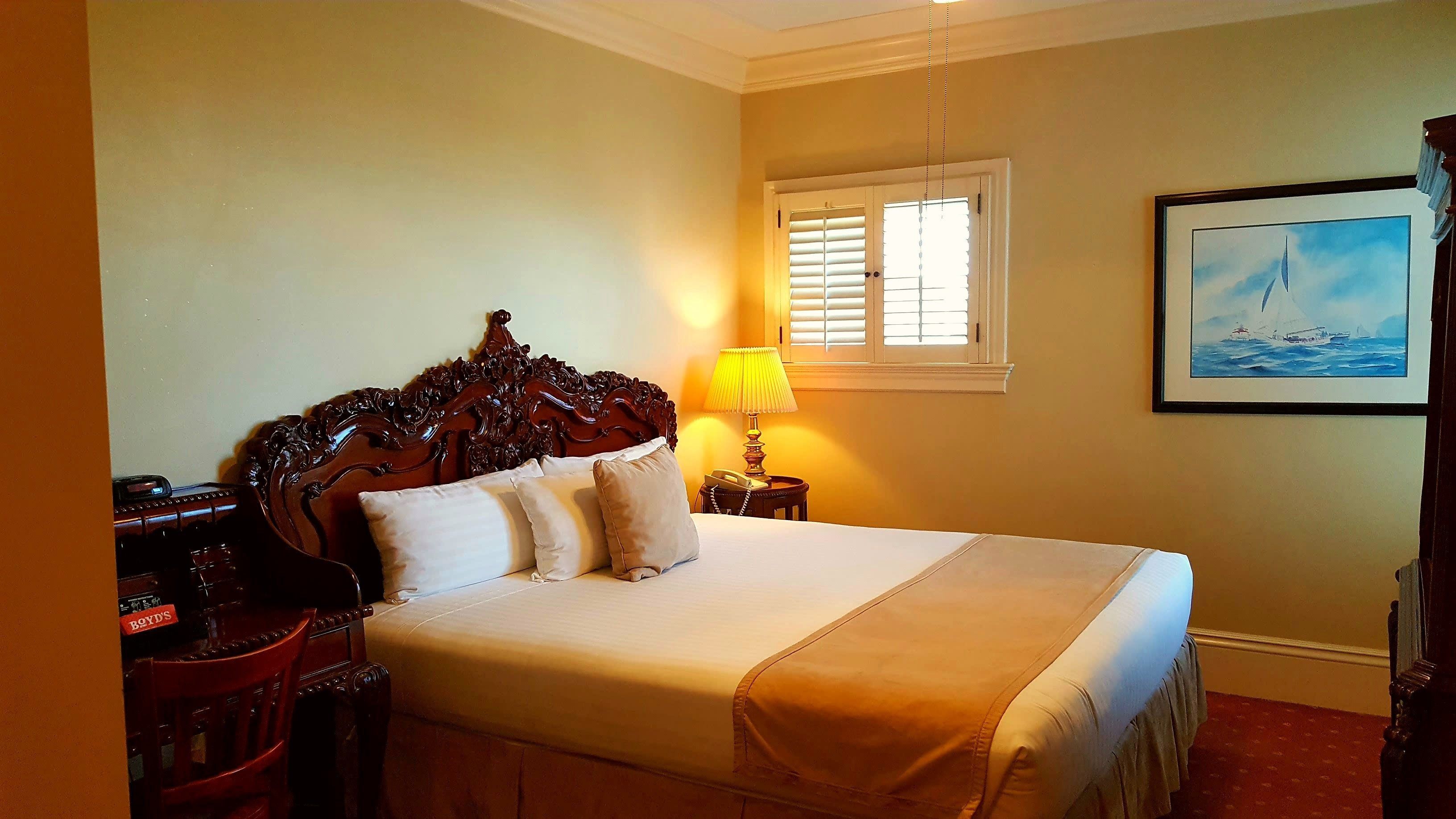 A hotel room with a large bed, a wooden headboard, a bedside table with a lamp, and a painting on the wall.