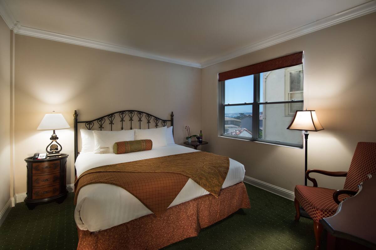 A neatly made bed with a patterned bedspread, a nightstand with a lamp, a chair, and a window with a view of buildings.