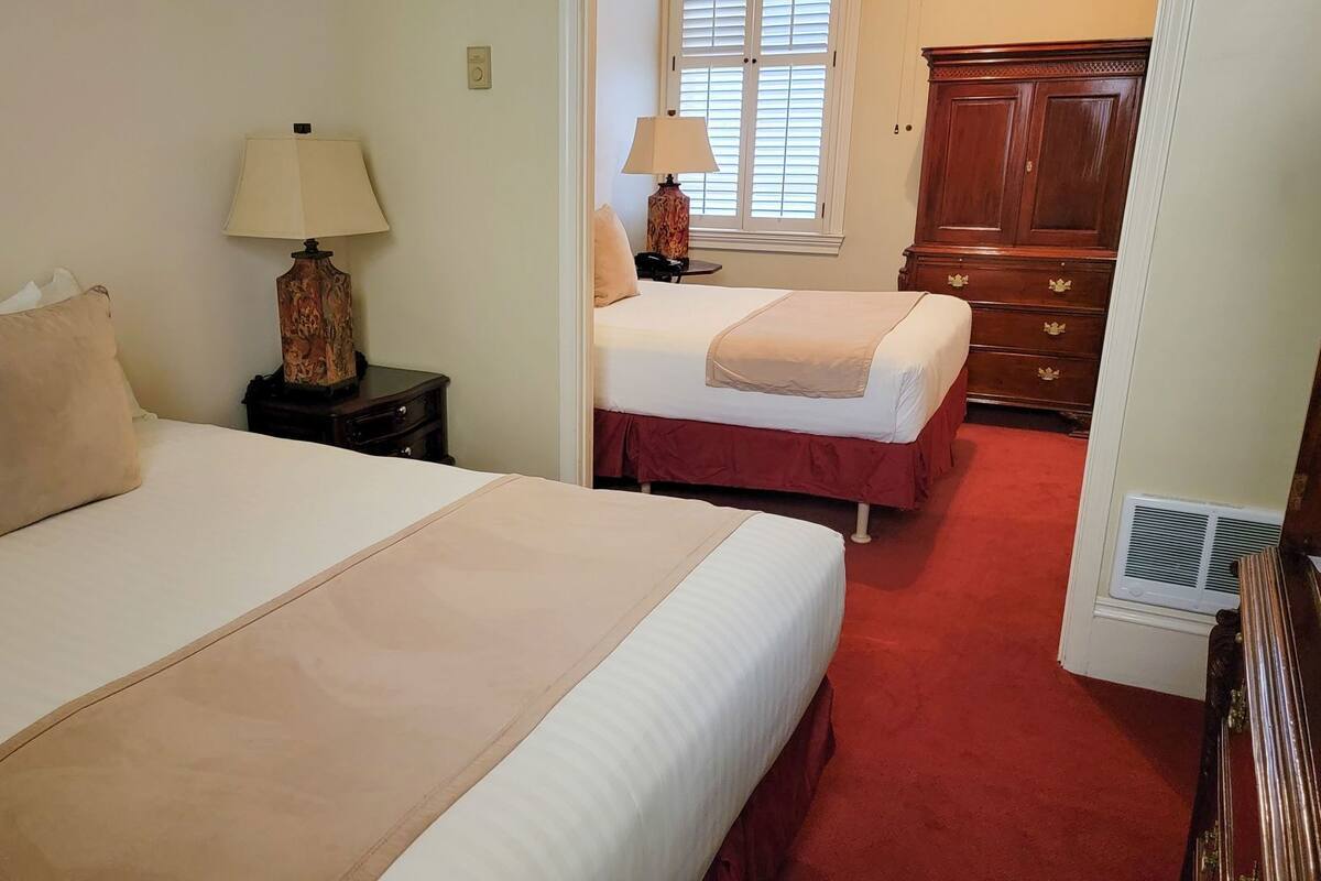 A hotel room with two beds, a nightstand with a lamp, a dresser, and a window with blinds.