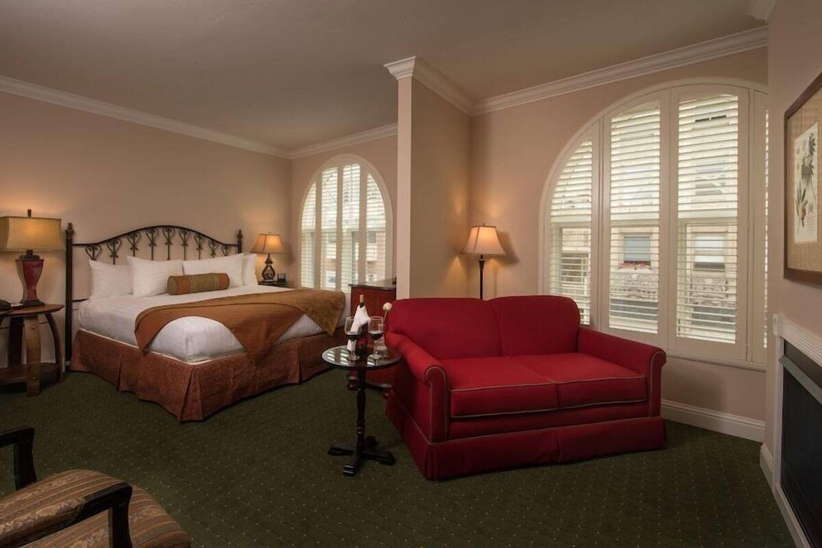 A hotel room with a bed, a red sofa, two lamps, a small table with a bottle and glass, and a framed picture on the wall.