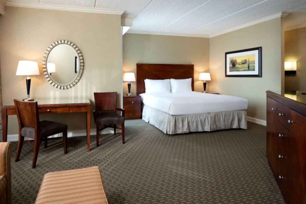 A hotel room with a bed, a desk, two chairs, a dresser, a mirror, and a painting on the wall.
