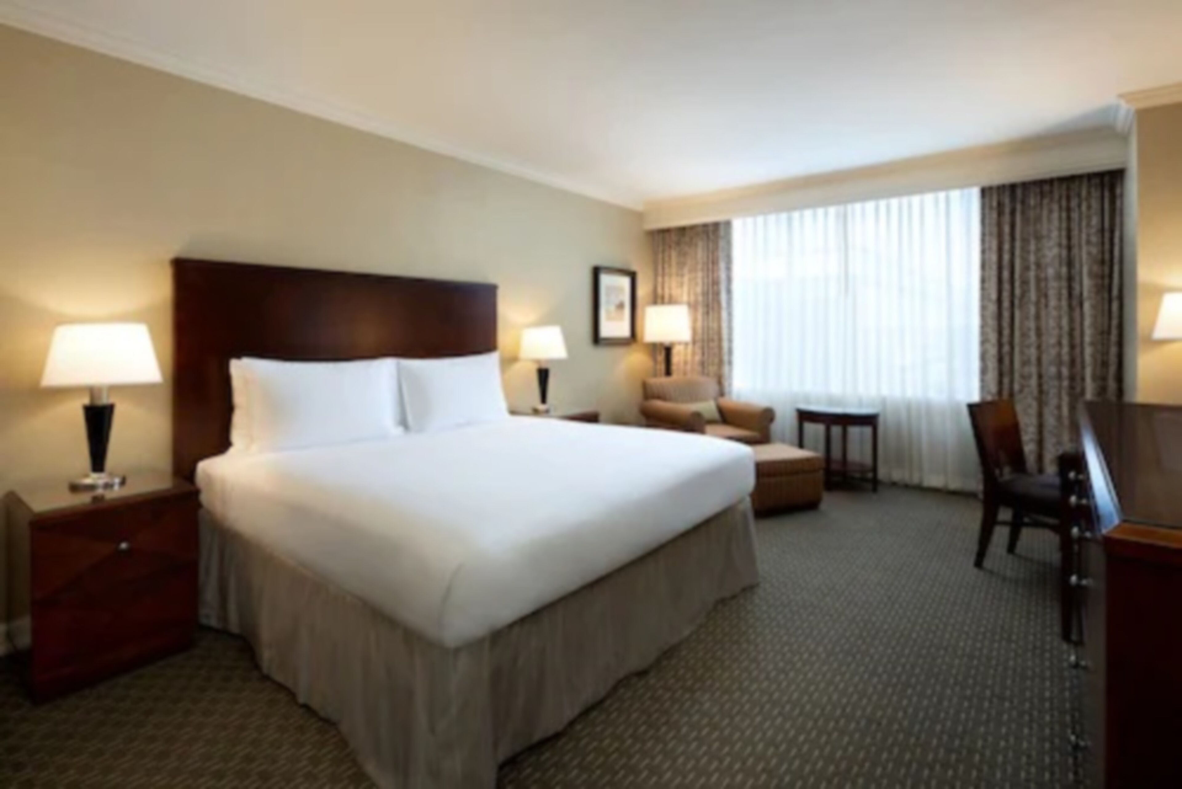 A hotel room with a large bed, two bedside tables with lamps, a small armchair, a desk, and a window with curtains.