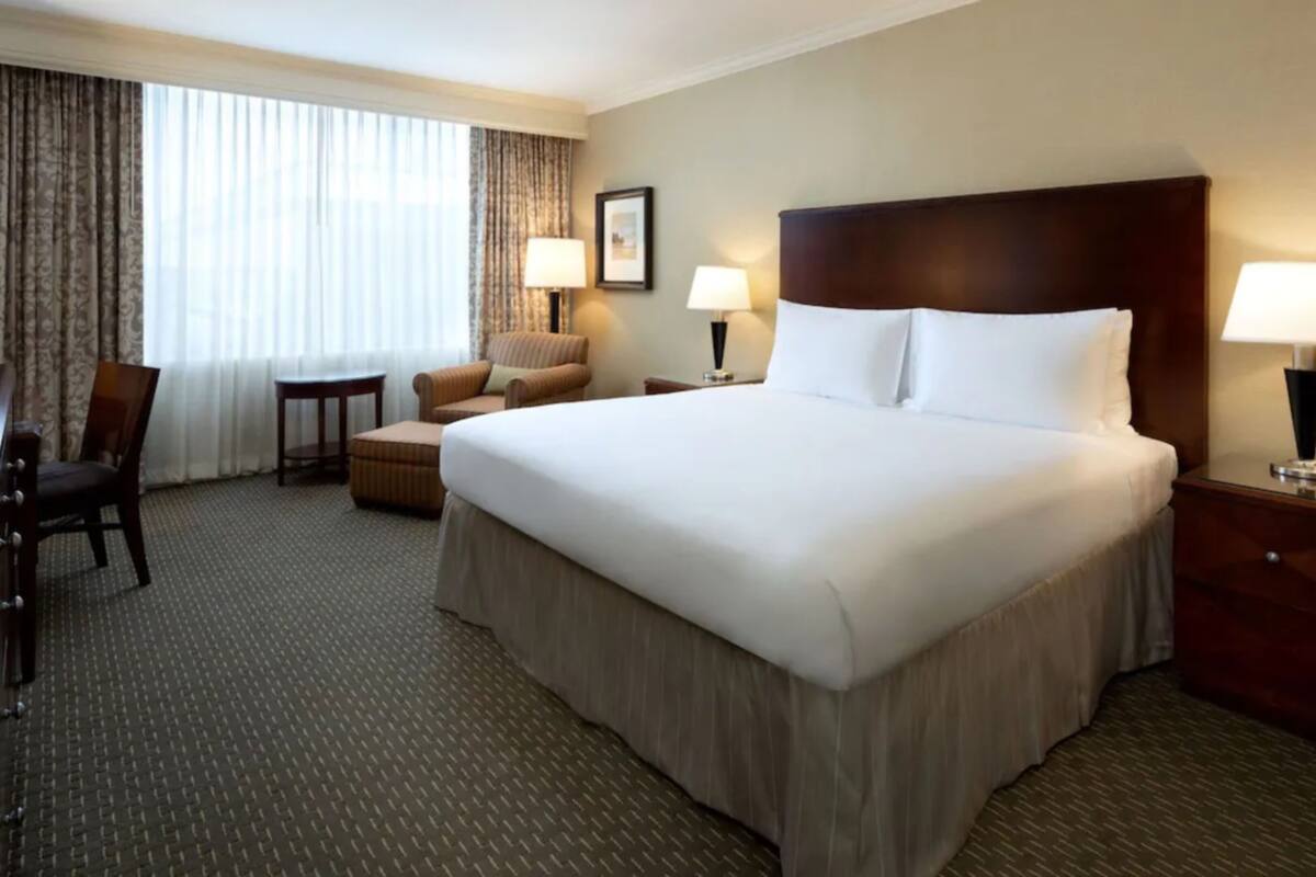 A hotel room with a large bed, two bedside tables with lamps, a chair, a small table, and a desk with a chair.