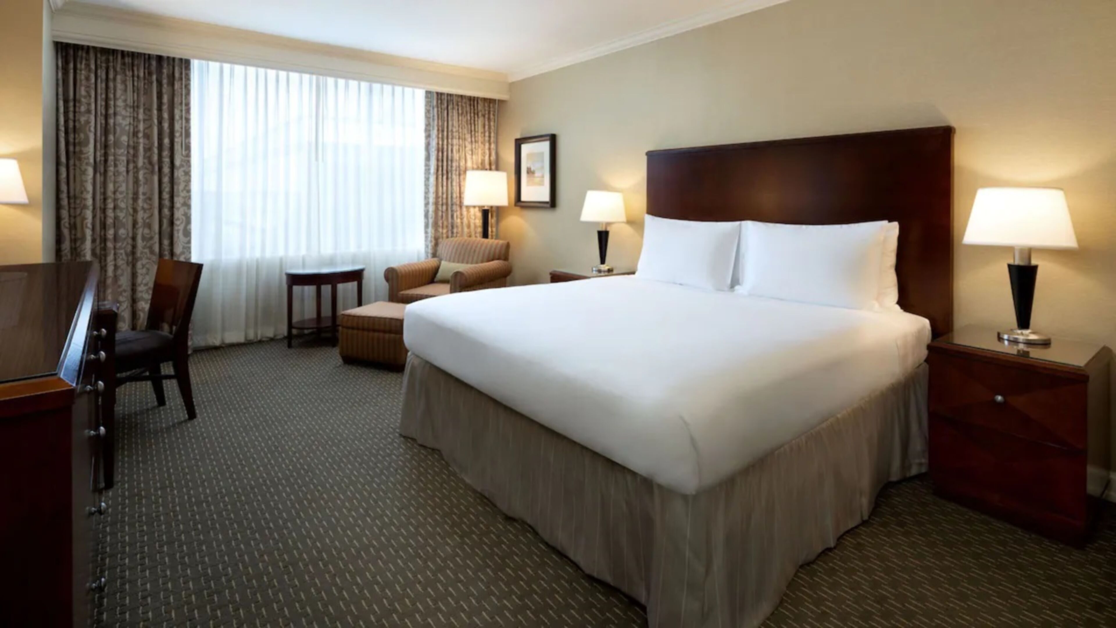 Une chambre d’hôtel avec un grand lit, deux tables de chevet avec des lampes, une chaise, une petite table et un bureau avec une chaise.