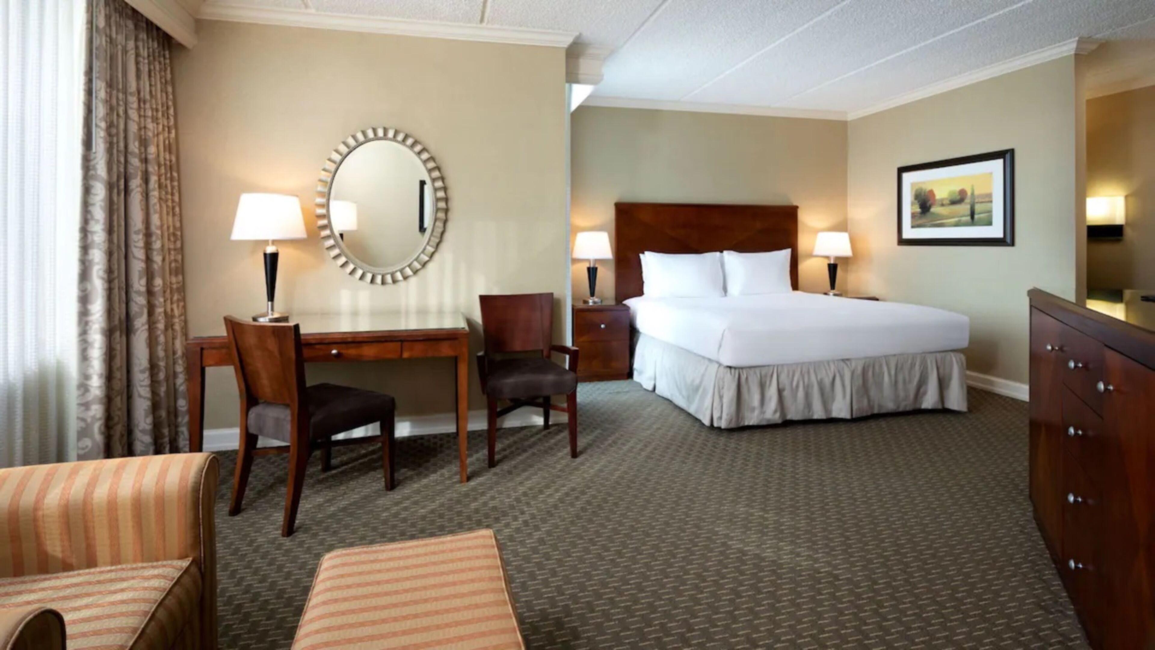 Une chambre d’hôtel avec un lit, un bureau, deux chaises, une commode, un miroir et un tableau au mur.