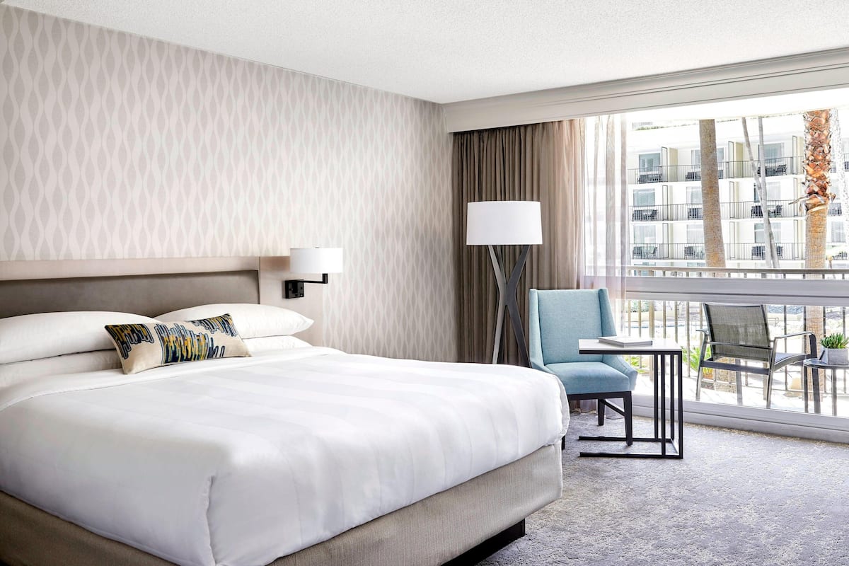 Ein Hotelzimmer mit einem großen Bett, einem Schreibtisch mit Lampe, einem Sessel und Blick auf das Gebäude gegenüber.