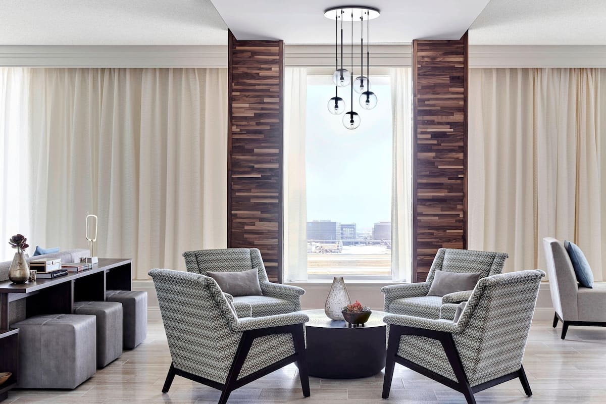 Ein modernes Wohnzimmer mit zwei Sesseln, einem Couchtisch und einem Beistelltisch. Es gibt ein großes Fenster mit Vorhängen, eine Wandpaneele aus Holz und eine Pendelleuchte.