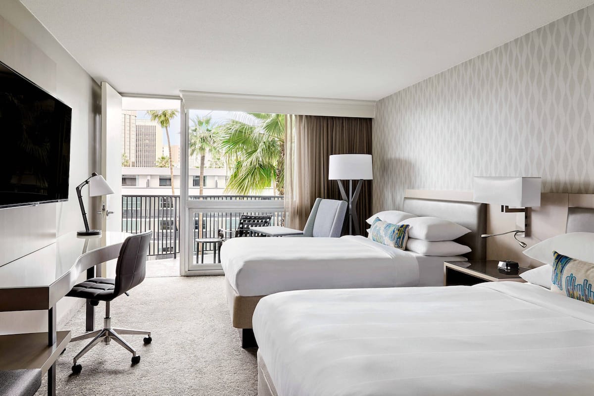 Ein Hotelzimmer mit zwei Betten, einem Schreibtisch, einem Sessel, einem Fernseher und einem Balkon mit Blick auf Palmen und Gebäude.