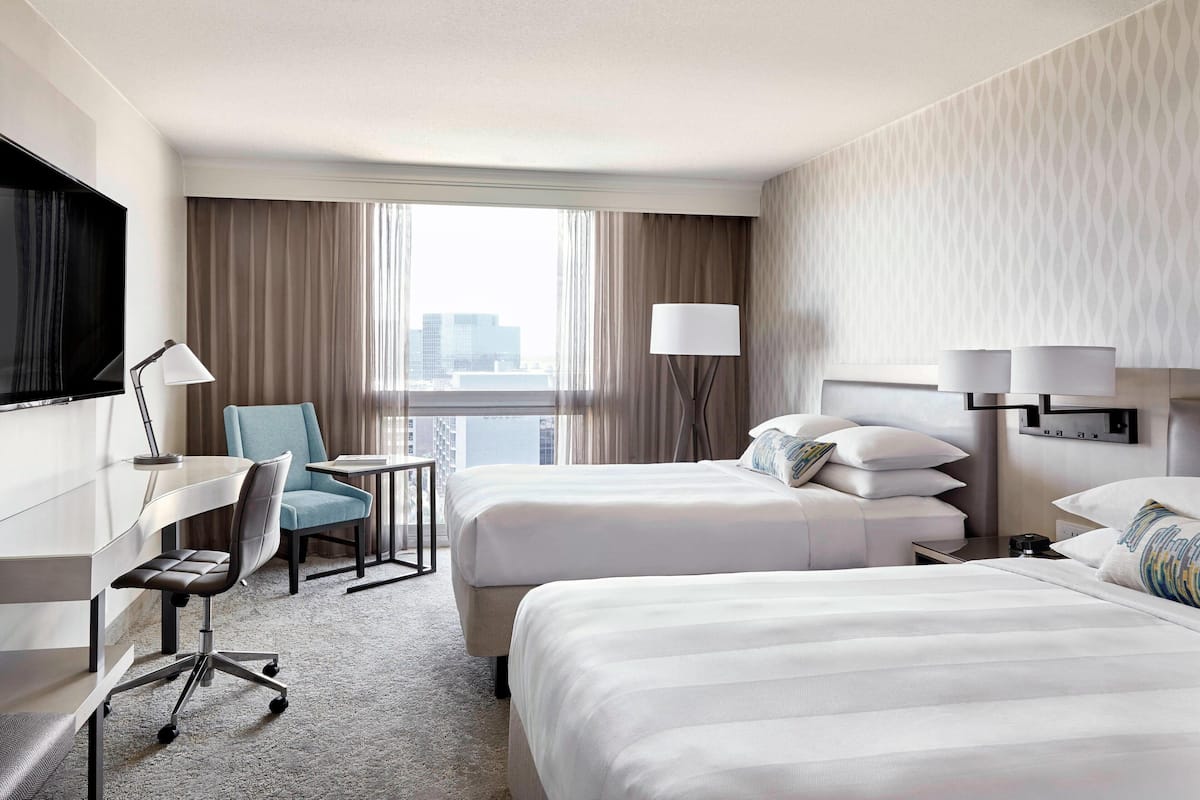 Ein Hotelzimmer mit zwei Betten, einem Schreibtisch, einem Stuhl, einem Fernseher und Blick auf die Stadt.