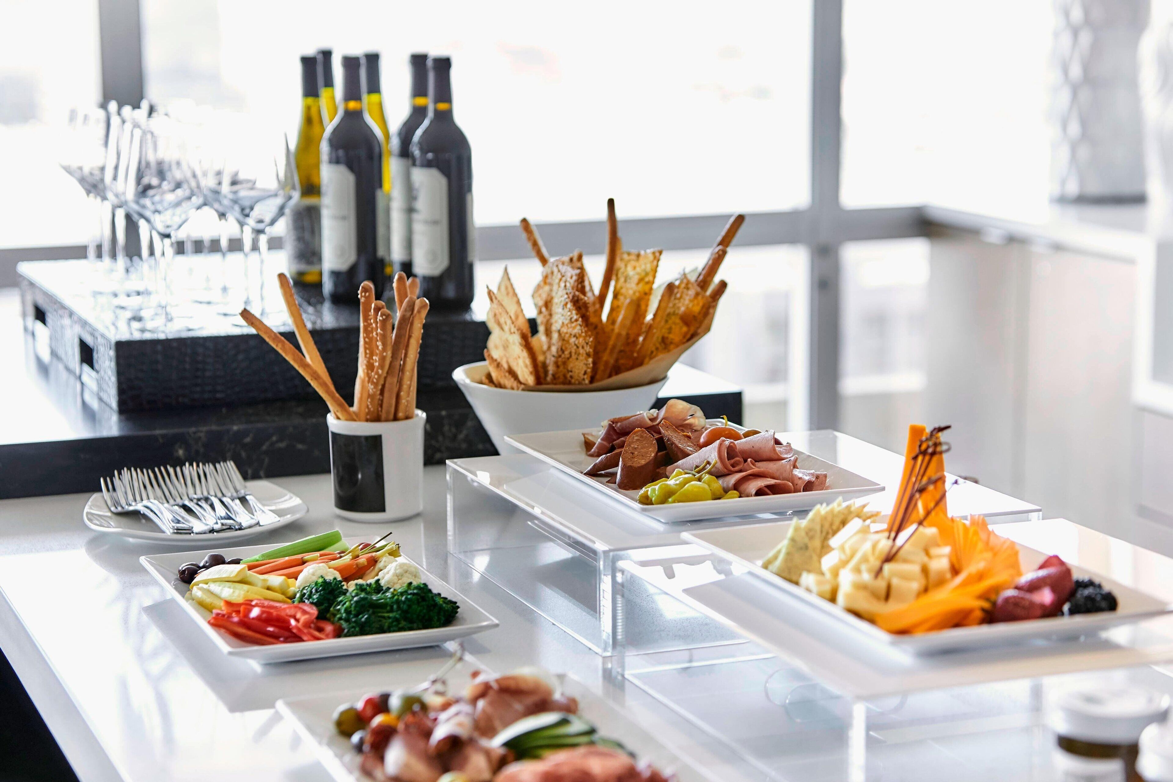 Una mesa de bufé con diversos alimentos, como frutas, verduras y carnes, junto con botellas de vino y copas.