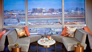 Un salon moderne avec des canapés gris, une table basse en verre et des tables d’appoint en cuivre. Une grande fenêtre offre une vue sur un aéroport avec de nombreuses pistes et des avions.
