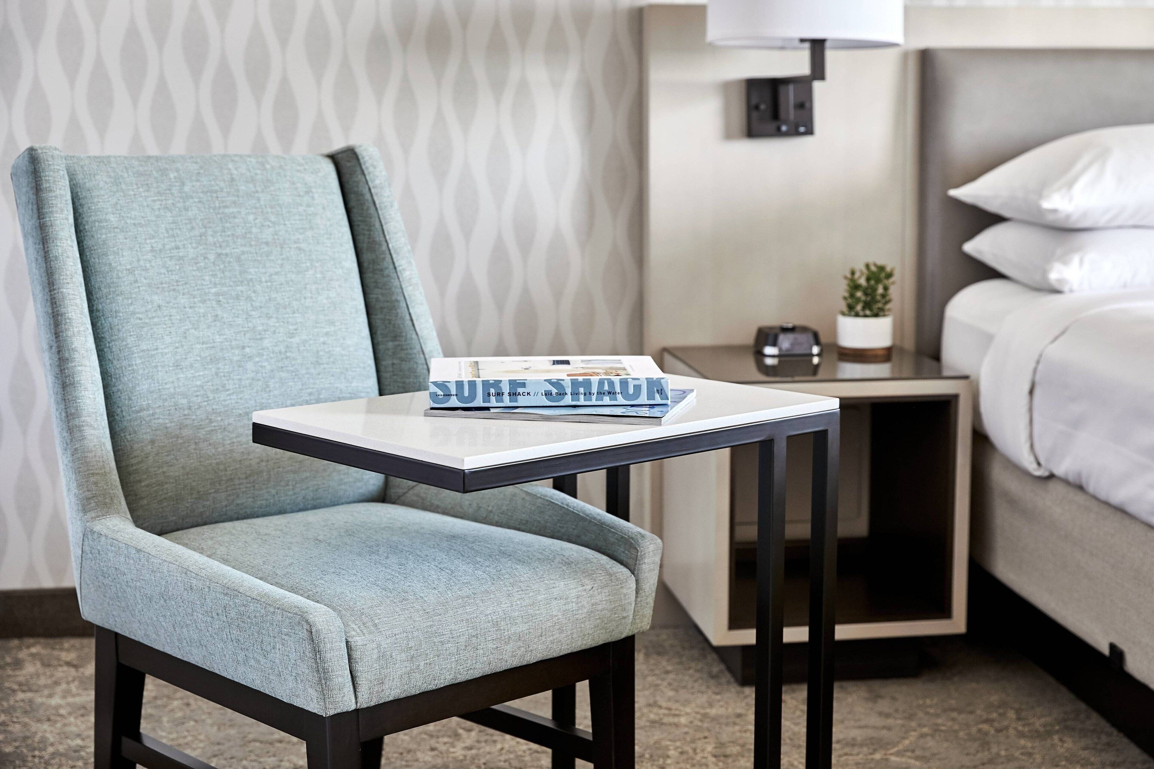 Habitación de hotel con un sillón azul claro, una mesita con una revista, una mesita de noche con una lámpara y una cama con ropa de cama blanca.