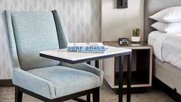 Habitación de hotel con un sillón azul claro, una mesita con una revista, una mesita de noche con una lámpara y una cama con ropa de cama blanca.