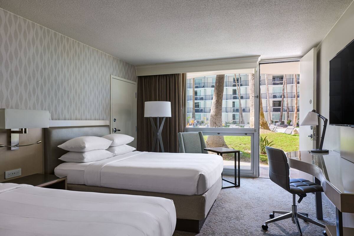 Ein Hotelzimmer mit zwei Betten, einem Schreibtisch, einem Stuhl, einer Lampe und Blick auf ein Gebäude sowie Grünfläche.