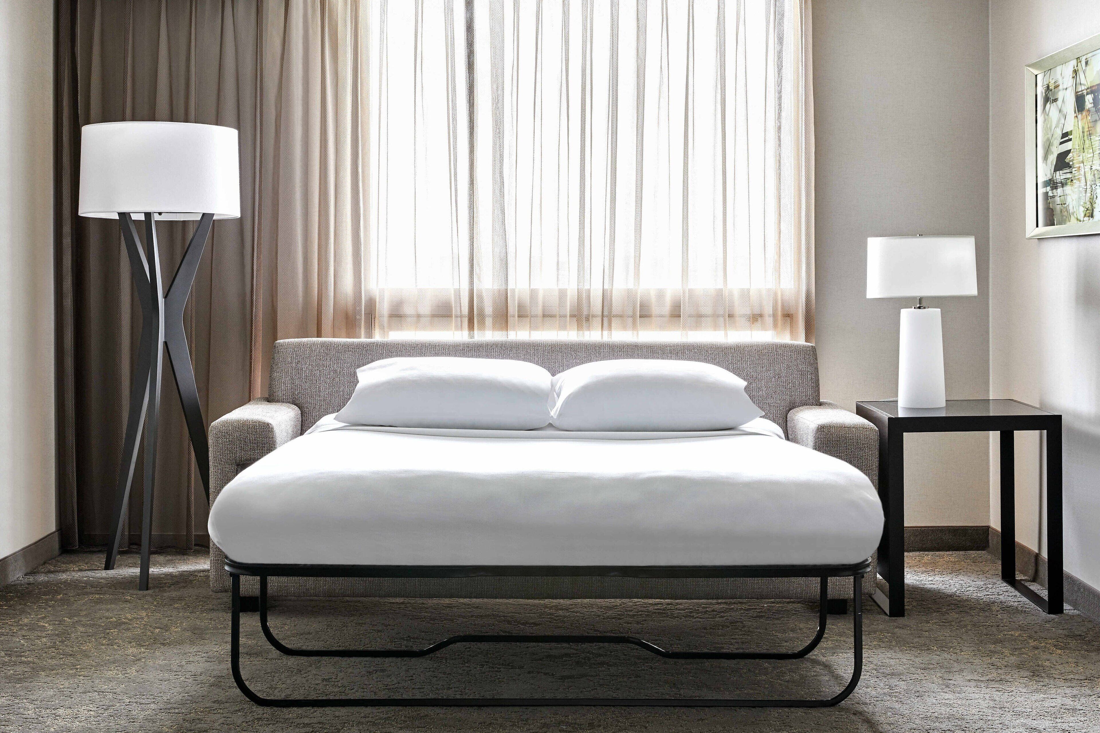Una habitación de hotel moderna con cama, lámpara de pie, mesita de noche y vistas a través de cortinas translúcidas.