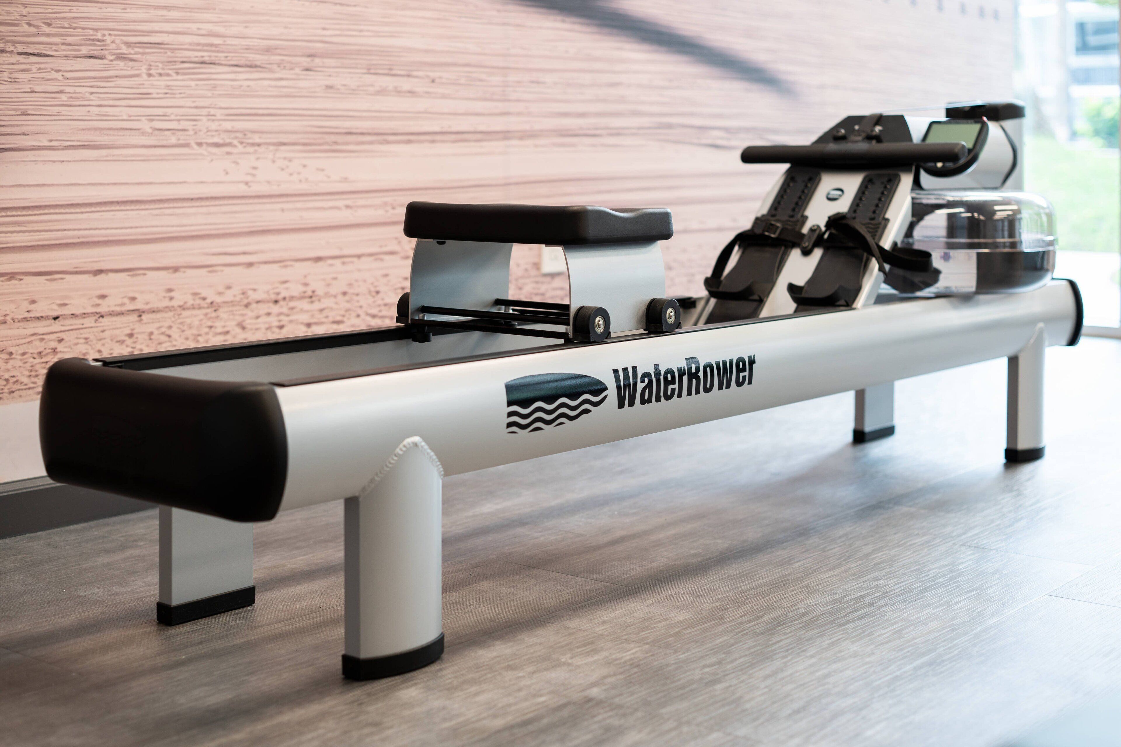 A stationary rowing machine branded "WaterRower" in a gym with a wooden floor and a plain wall in the background.