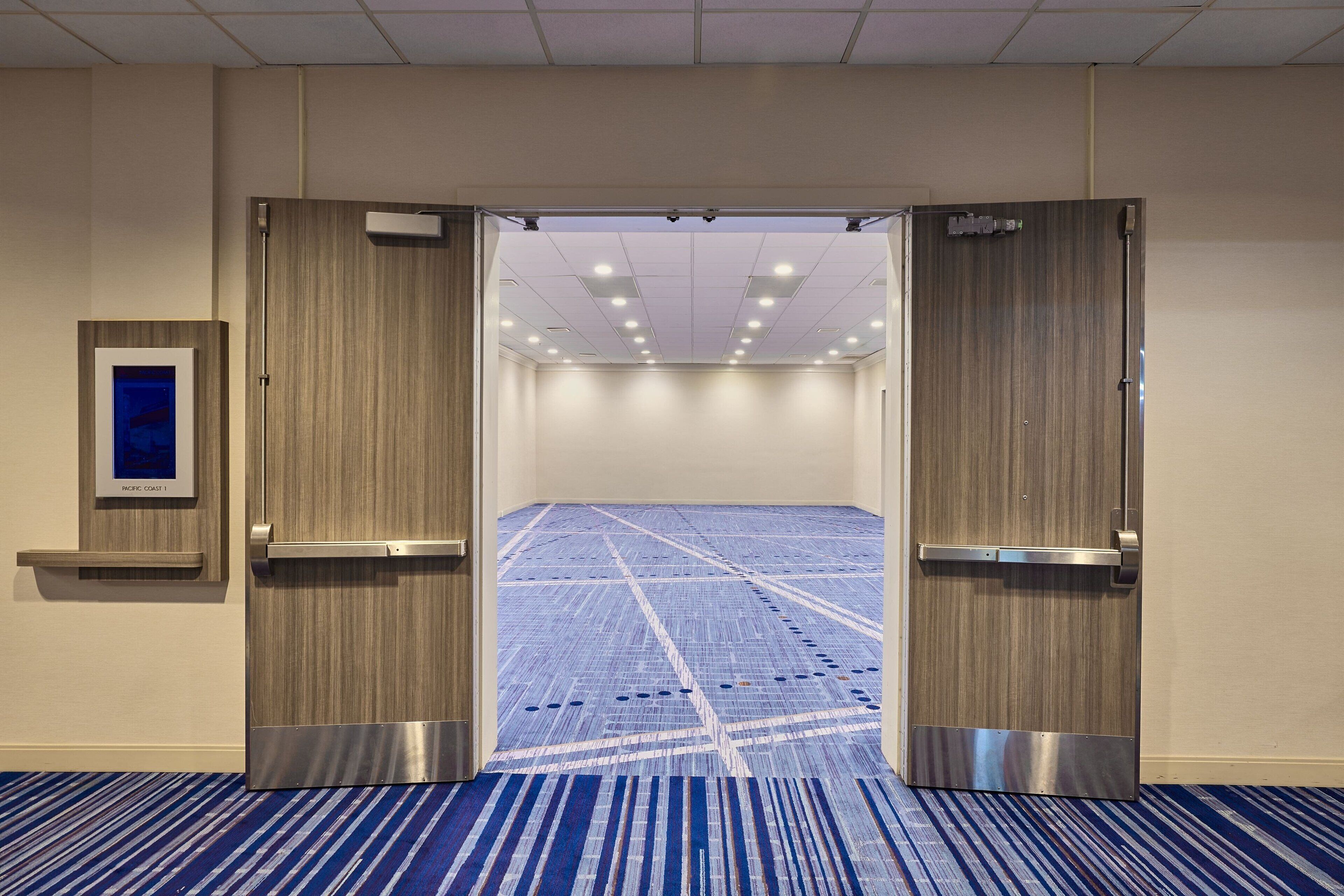 Entrada de una habitación de hotel con alfombra azul, un cuadro enmarcado en la pared e iluminación empotrada en el techo.