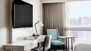A hotel room with a flat-screen TV mounted on the wall, a desk with a chair, a small table, and a bed.