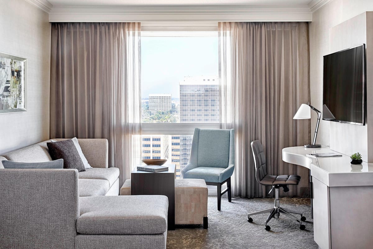 Ein modernes Hotelzimmer mit Sofa, Sessel, Schreibtisch und einem großen Fenster mit Vorhängen.