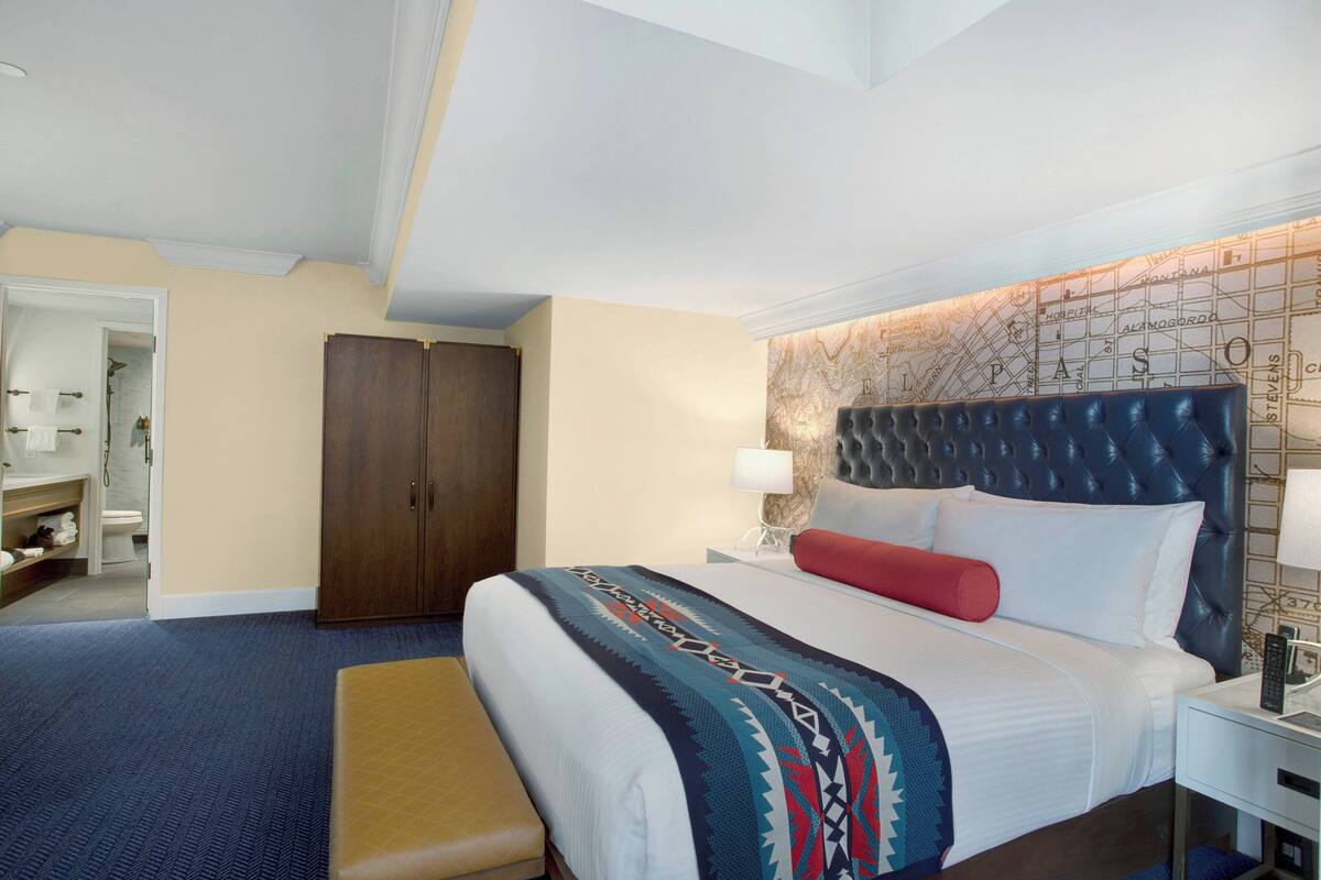 A hotel room with a large bed, a wooden wardrobe, a bench, and a map-themed headboard.