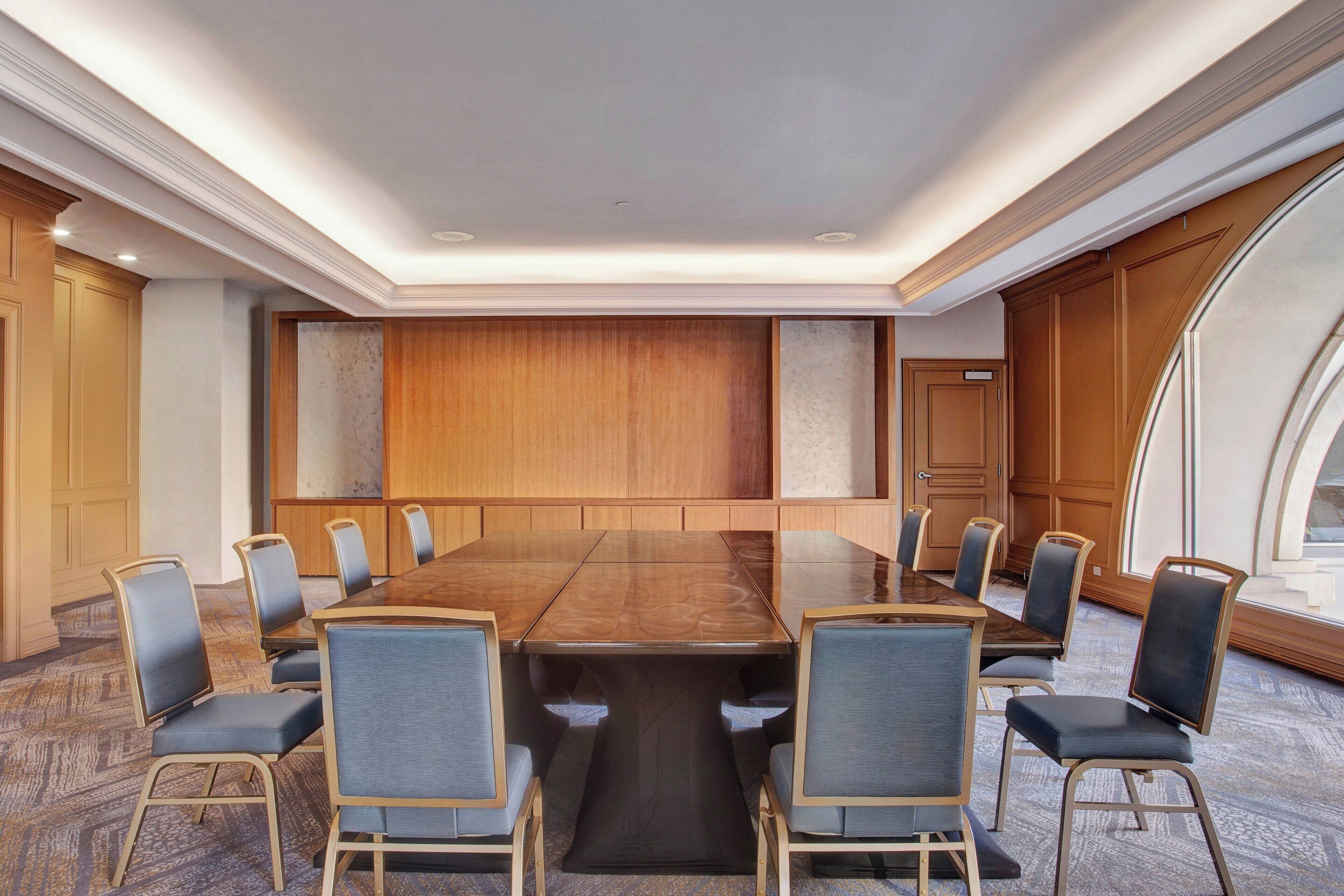 A conference room with a long table and 10 chairs, wooden paneling on the walls, and a large window on the right.