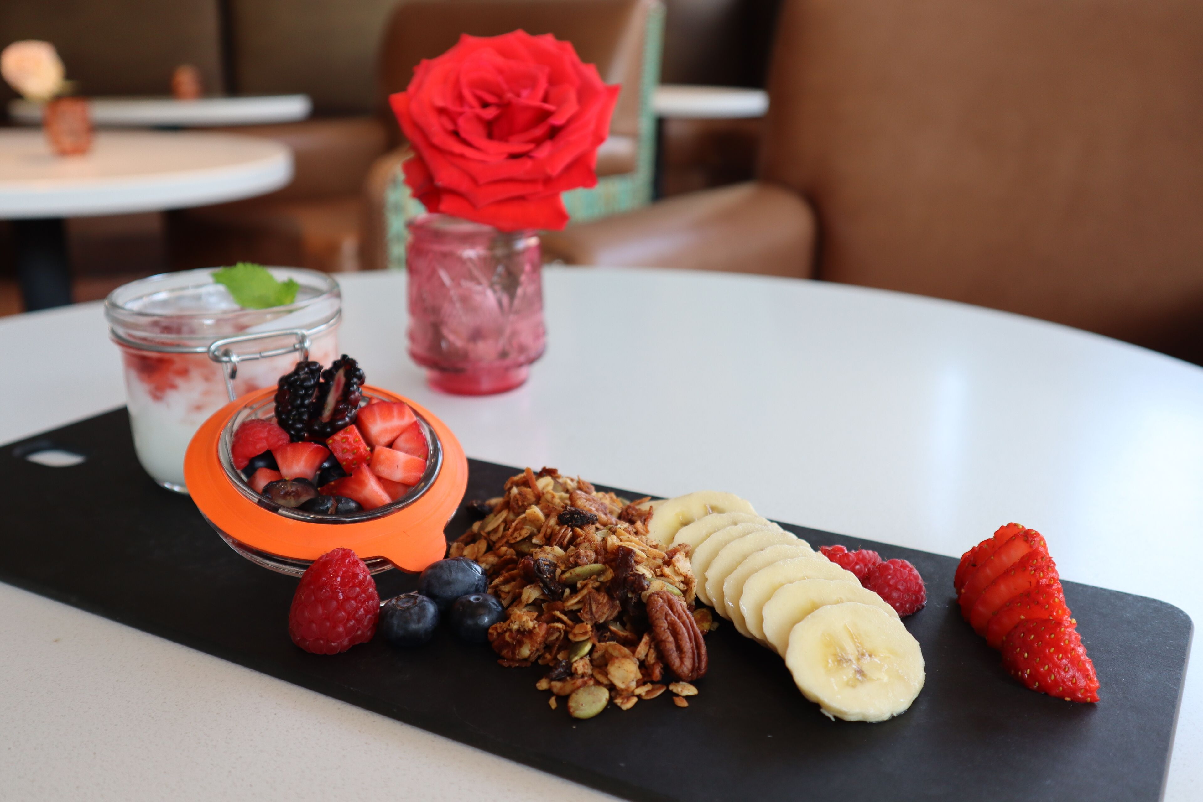 Un plateau de petit-déjeuner avec du yaourt, du granola, des fruits rouges et une rose dans un bocal en verre.
