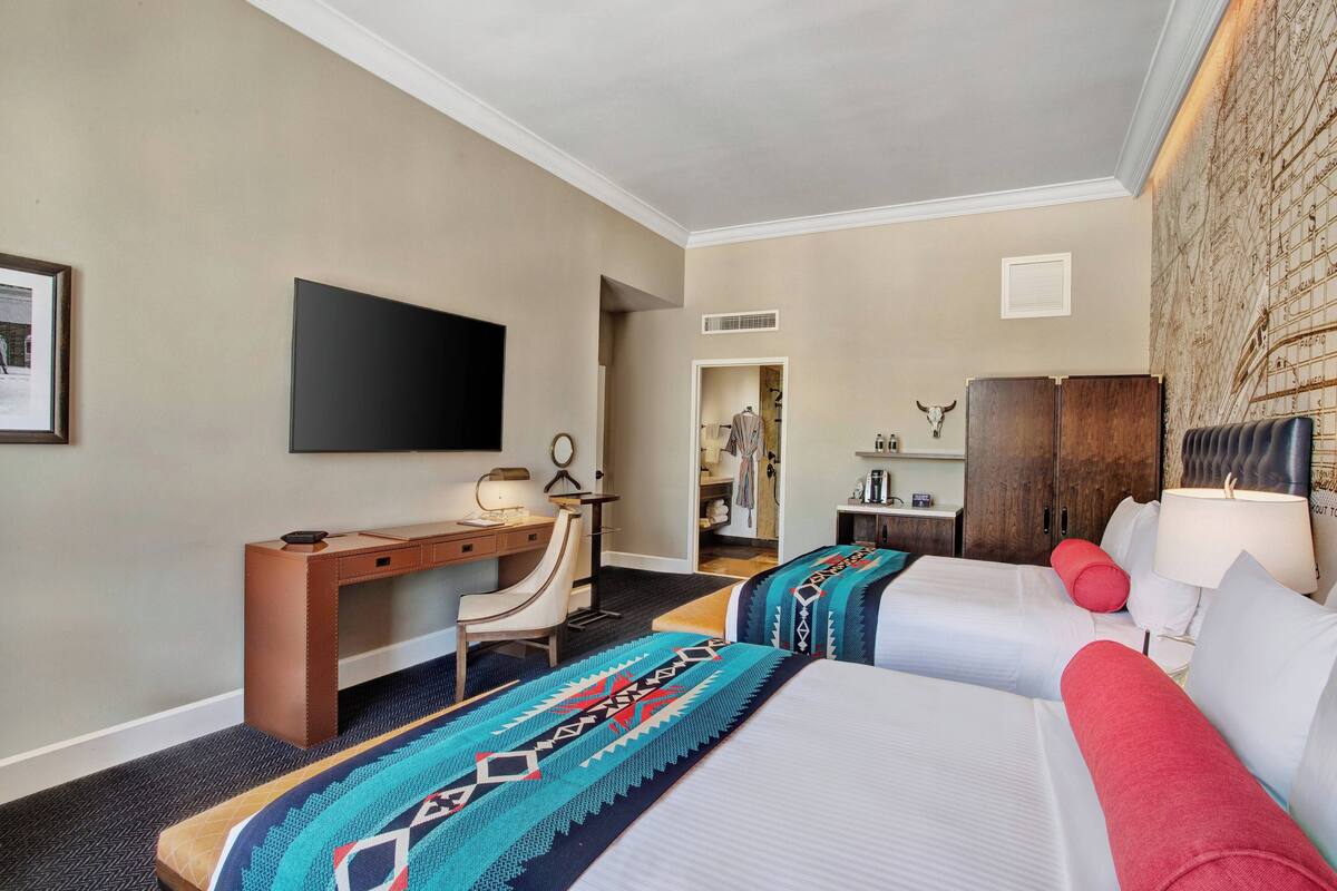 A hotel room with two beds, a desk, a chair, a TV, and a bathroom visible through an open door.