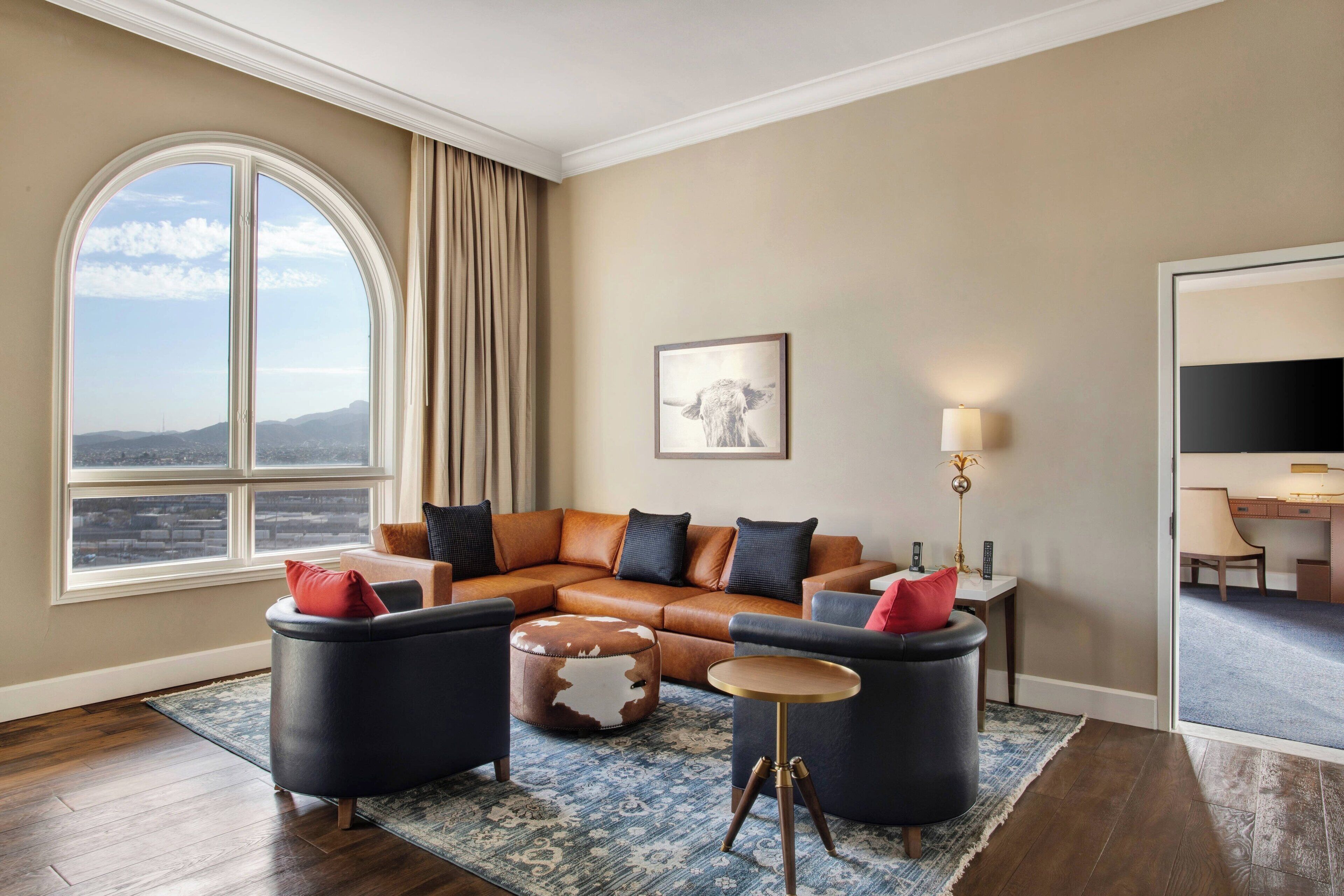 A modern living room with a large window, a leather sofa, two armchairs, a round coffee table, and a flat-screen TV.