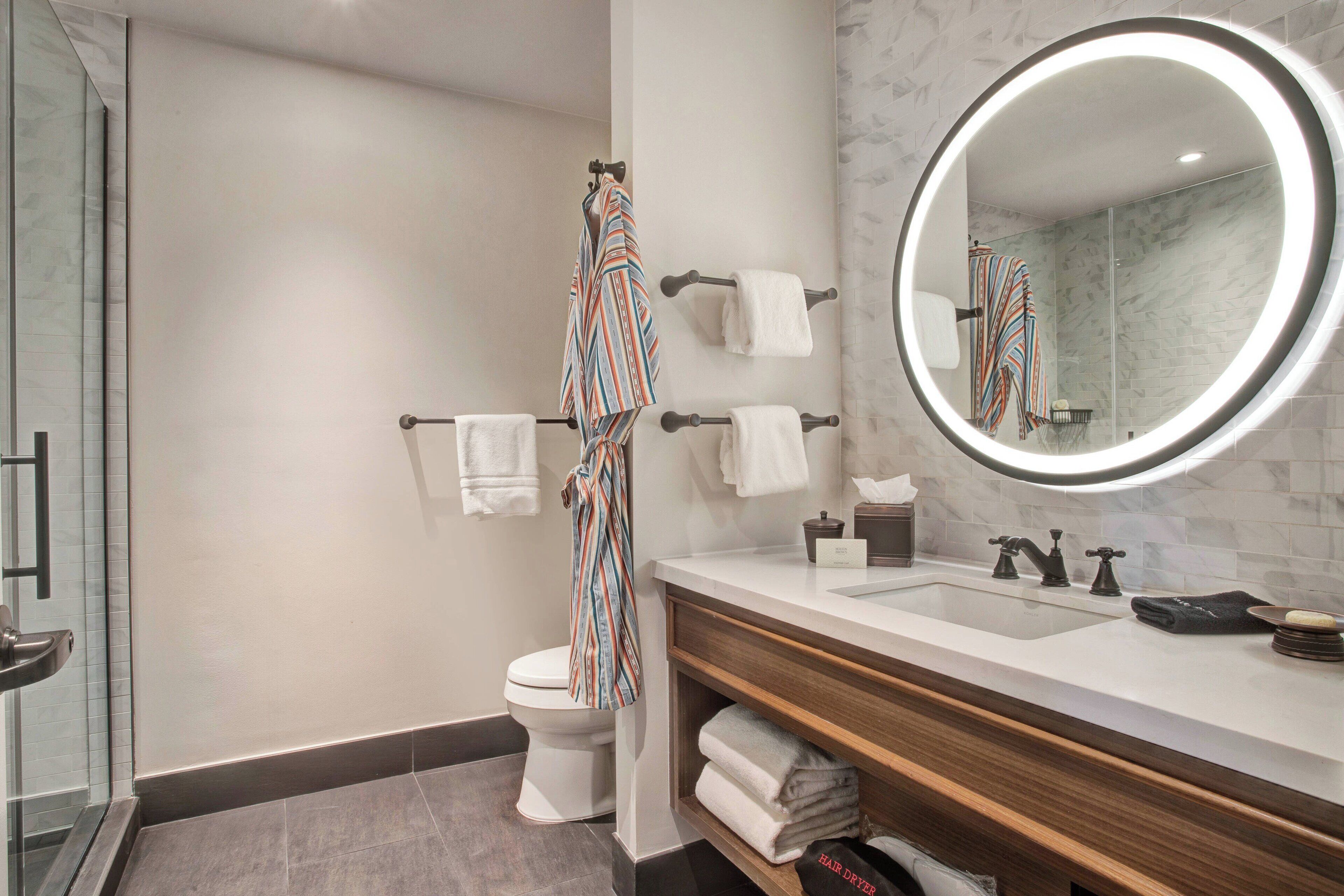 A modern bathroom with a white toilet, a large mirror with a circular light, a wooden vanity, and a glass shower enclosure.