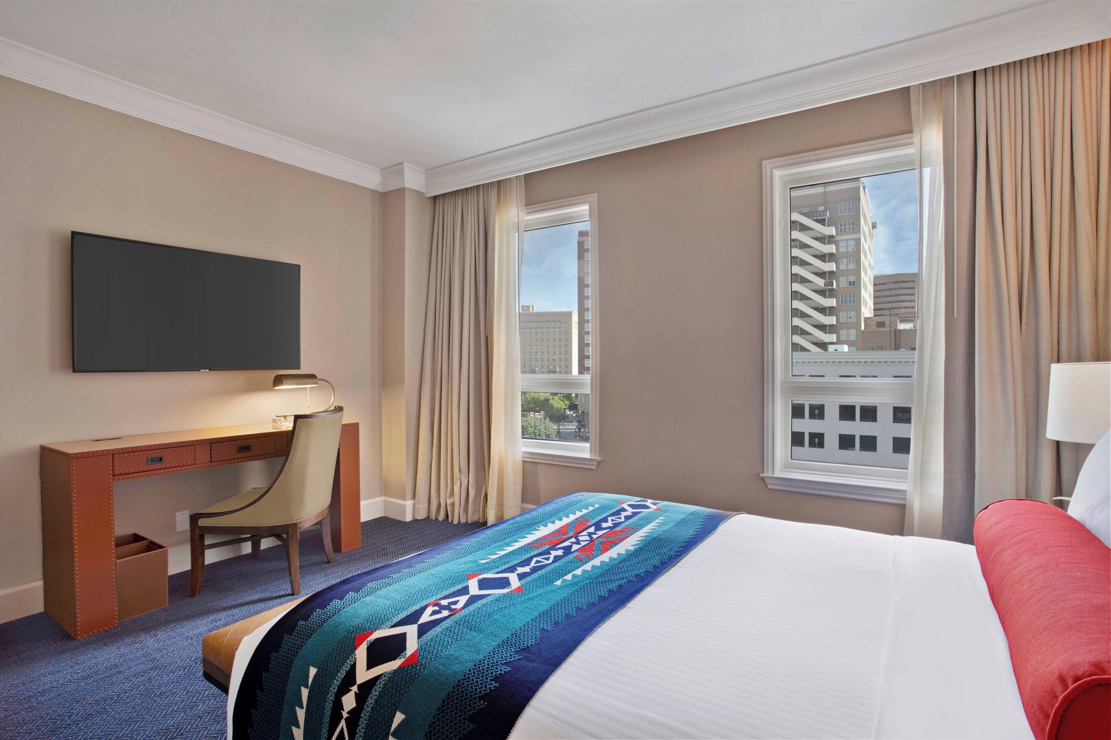A hotel room with a bed, a desk, a chair, a television, and large windows.