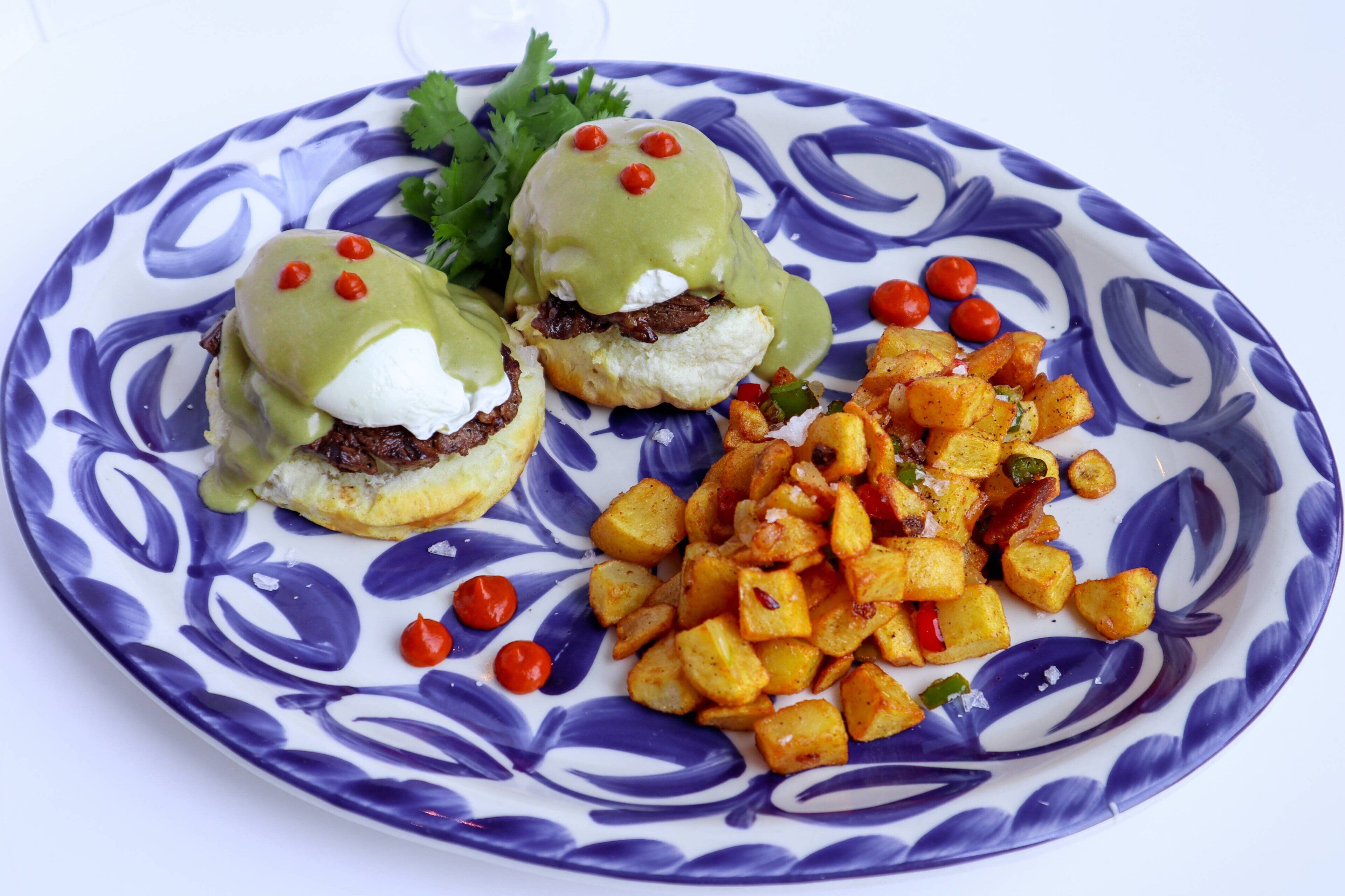 Un plato con dos sándwiches cubiertos de salsa verde y pimientos rojos, acompañado de una guarnición de patatas asadas.