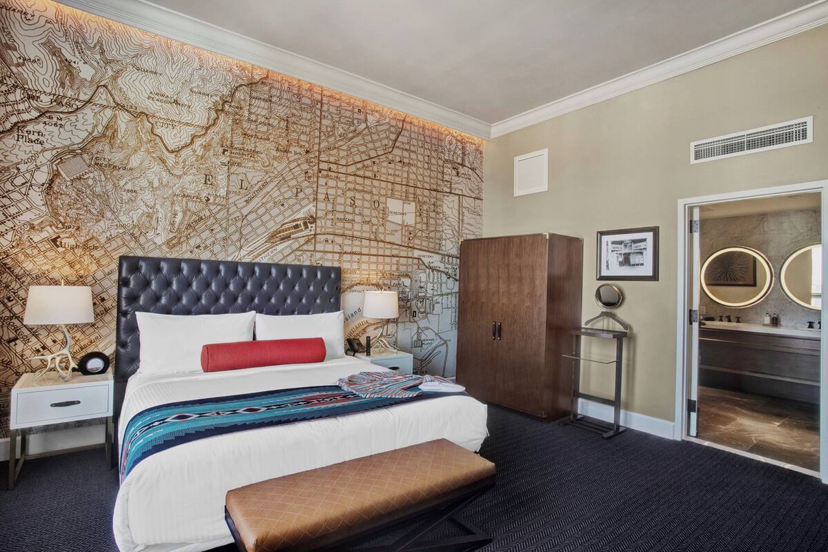 A hotel room with a large bed, a map wall, a wardrobe, a vanity with mirrors, and a bench.